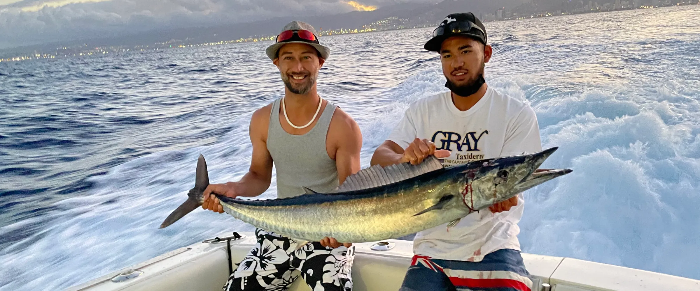 Two men on a boat proudly holding a large fish with the ocean and mountains in the background at sunset.